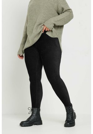 Osoba nosząca czarne botki i czarne legginsy oraz szary, teksturowany, oversize'owy sweter, stojąca na tle jednolitego, jasnego tła.