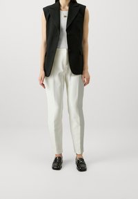 Black sleeveless vest over a white ribbed top, paired with white tapered pants and black loafers. Features sharp lapels and two buttons.