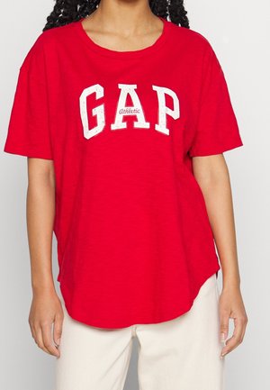 Person iført en løs rød t-shirt med "GAP Athletic" i hvide bogstaver, kombineret med lyse bukser mod en ensfarvet baggrund.