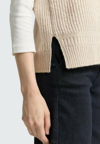 Beige gebreide trui met een ribbelstructuur en zijsplitten, gecombineerd met donkerblauwe jeans. Close-up van een hand die op de jeans rust.