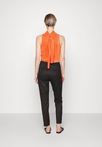 Pomarańczowy plisowany top bez rękawów z wiązaniem przy szyi, zestawiony z dopasowanymi czarnymi spodniami i płaskimi sandałami, widziany od tyłu.