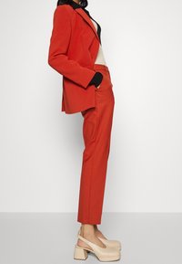 Czerwona, dopasowana marynarka i spodnie o prostych nogawkach w tym samym kolorze, zestawione z beżowymi butami na platformie. Marynarka ma klapy z wcięciem i kieszenie.