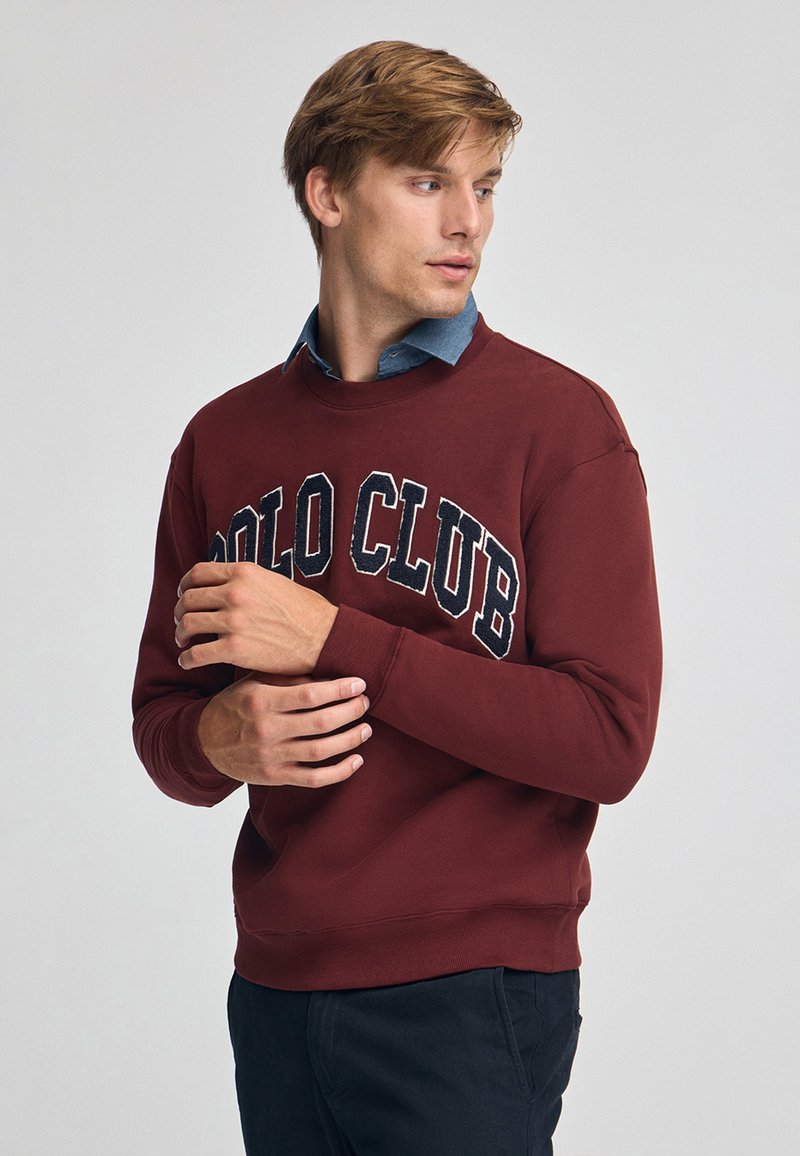 Vinröd sweatshirt med "POLO CLUB" broderat över bröstet, ribbade mudd och nederkant, bärs över en blå skjorta med krage. Slät textur.