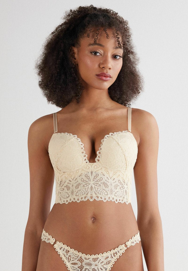 Spetsunderkläder i mjuk beige med en djup V-ringad bralette med vågiga kanter och tunna axelband, kompletterat med matchande spetsunderbyxor.