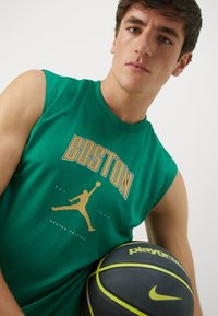Green sleeveless shirt featuring "BOSTON" in gold above a jumping figure graphic, paired with a black basketball marked with yellow accents.