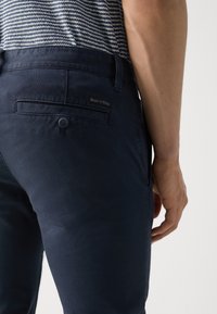 Chino in cotone blu navy con una texture liscia, dotati di una tasca posteriore con bottone e un piccolo tag con il marchio, mostrati di lato.