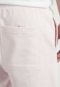 Un pantalon de survêtement rose clair avec une poche arrière dotée d'un surpiqure saisissante, fabriqué en tissu doux et texturé. Ceinture élastique visible.