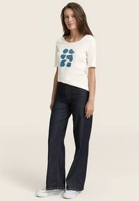 White short-sleeve t-shirt with blue abstract design, paired with dark blue flared jeans and white sneakers. Smooth fabric texture.