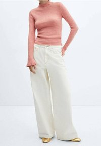 Woman wearing a fitted pink turtleneck sweater, wide-leg white pants, and gold pointed shoes, standing against a plain white background.