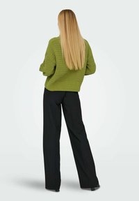 Groene gebreide trui met een gestructureerd patroon, oversized pasvorm en geribbelde zoom, gecombineerd met zwarte wijde broek. Lange, steile blonde haren zichtbaar.