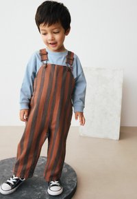Stribede brune og grå overalls med metalspænder, parret med en lys blå langærmet skjorte og sorte sneakers. Glat stof, afslappet pasform.