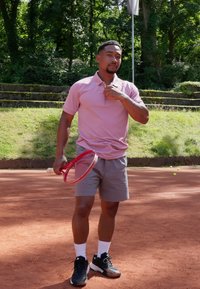 Jugador de tenis con una camiseta polo rosa clara y pantalones cortos grises, sosteniendo una raqueta roja en una cancha de arcilla, con zapatillas deportivas negras y calcetines blancos.