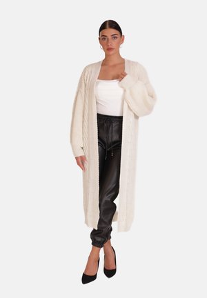 Lunga giacca a maglia crema con motivo a treccia, indossata sopra una maglietta bianca e pantaloni in pelle nera, con maniche larghe e apertura frontale.