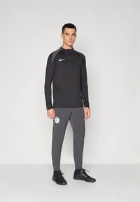 Muž oblečený v čiernom tričku s dlhými rukávmi Nike a sivých nohaviciach s logom PSG, so španielskymi topánkami, stojaci pred jednoduchým svetlým pozadím.