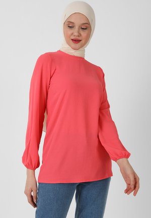 Coral long-sleeve top with a rounded neckline and gathered cuffs, paired with light blue denim jeans; soft fabric with a smooth texture.
