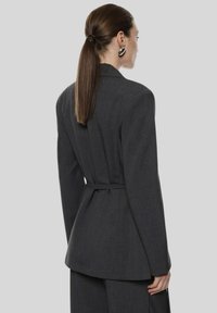 Donna con capelli marroni lisci raccolti in una coda di cavallo bassa, che indossa un blazer grigio scuro con cintura e pantaloni abbinati, rivolta di profilo su uno sfondo grigio.