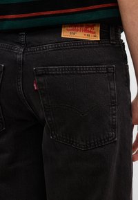 Κοντινό πλάνο ενός ατόμου που φοράει μαύρα τζιν Levi's 578 με ορατή πίσω τσέπη, κόκκινη ταμπέλα Levi's και δερμάτινη ετικέτα σε ταμπά χρώμα που δείχνει το μέγεθος W31 L34.