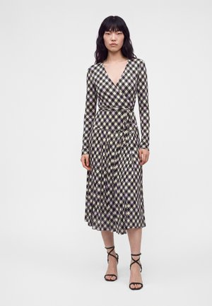 Femme portant une robe portefeuille midi à manches longues avec un col en V et un motif à carreaux noir, blanc et violet clair, associée à des talons noirs à brides.