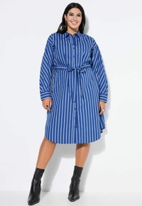 Robe à boutons rayée bleue avec des manches longues et une taille nouée. Le tissu semble lisse, associé à des bottines noires élégantes.