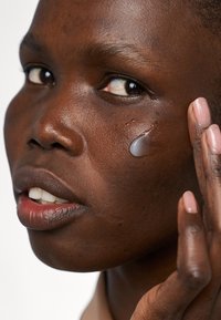 En klar gel appliceras på ansiktet, vilket framhäver den släta, mörka huden. Personen har kort hår och neutral nagellack, med ett fokuserat uttryck.