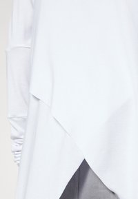 Tunic bianca a maniche lunghe con orlo asimmetrico, caratterizzata da un design a strati e un tessuto morbido e liscio. Dettagli testurizzati sulle maniche.