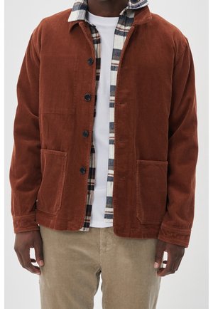 Mand iført en rustfarvet kordfløjsjakke over en hvid og ternet skjorte, kombineret med beige kordfløjsbukser, vist fra nakke til hofter.