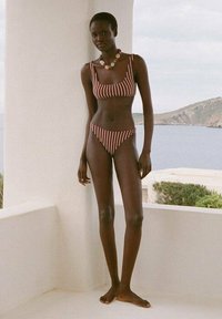 Bikini a due pezzi a righe in borgogna e crema. Il reggiseno ha spalline larghe, mentre la parte inferiore presenta un taglio a vita bassa. Una collana colorata di perline completa il look.