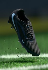 Sorte fodboldstøvler med struktureret overdel, lyseblå accenter og perforeringer for åndbarhed. Støvlerne vises på grøn græs med flyvende affald.