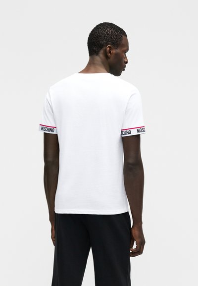 Άνδρας με γυρισμένη την πλάτη που φοράει ένα λευκό κοντομάνικο μπλουζάκι με την επιγραφή "MOSCHINO" στις άκρες των μανικιών και μαύρο παντελόνι.