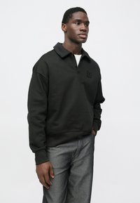 Melns pulovers ar salocītu apkakli, priekšējo pogu apdari un izšūtu logotipu. Modelis valkā ar tumšām džinsu biksēm. Gluda auduma tekstūra.