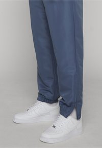 Κάτω άκρα με μπλε φόρμες joggers που έχουν ελαστικές μανσέτες και πλαϊνά φερμουάρ, συνδυασμένα με λευκά παπούτσια Nike Air Force 1 πάνω σε γκρι πάτωμα.