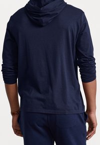 Muž v námořnickém modrém svetru s dlouhými rukávy a odpovídajících kalhotách, pohled zezadu s rukama u těla.