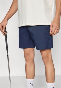 Σκούρα μπλε σορτς με λεία υφή, ίσια εφαρμογή και ραμμένες άκρες· σετ με λευκές κάλτσες αστραγάλου που έχουν μαύρο λογότυπο.