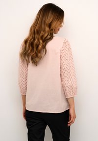 Rosa Bluse mit drei Viertelärmeln, die ein Chevron-Muster und Stickdetails aufweist, kombiniert mit schwarzen Hose. Weicher Stoffeindruck.