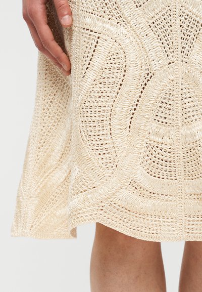 Main tenant une jupe beige au crochet avec des motifs circulaires et spiraux, montrant la texture et le détail du tricot près des jambes nues.