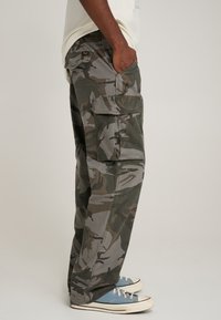 Cargo pants in a green and gray camouflage pattern, made of durable fabric with multiple pockets and a relaxed fit, paired with blue sneakers.