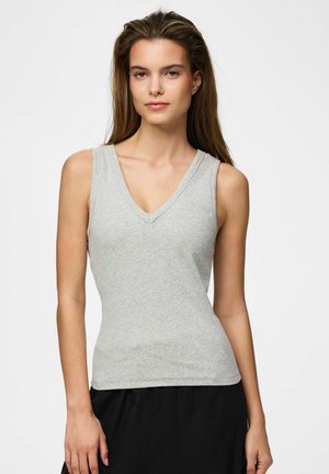 Tânără cu părul lung și brunet, purtând un top gri deschis fără mâneci cu decolteu în V și pantaloni negri, stând pe un fundal alb simplu.