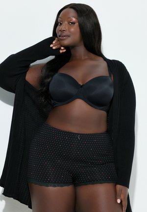 Schwarzer gepolsterter BH aus glattem Stoff, kombiniert mit schwarzen Shorts mit weißen Punkten, Spitzenabschluss und kleinem Schleifendetail. Im Hintergrund ein geknöpfter Cardigan.