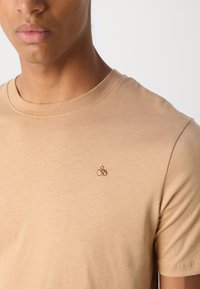 Camiseta de algodón beige con cuello redondo y un pequeño logotipo bordado marrón en el lado izquierdo del pecho. Textura suave, diseño ajustado.