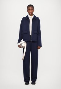 Navy blue jacket with two front pockets, paired with a light turtleneck and wide-leg navy trousers. Holding a small white bag with a ribbon.