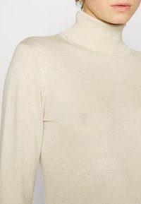 Beige Rollkragenpullover aus dünnem Rippstrick, mit hohem Kragen und taillierter Silhouette. Glatte Textur, minimalistische Details.