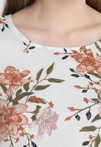 Blusa blanca de mujer con cuello redondo, decorada con un estampado floral en rojo, púrpura y naranja, acompañado de hojas verdes y pequeños capullos azul oscuro.