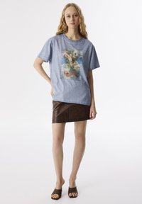 Mujer con largo cabello rubio que lleva una camiseta gráfica azul, una falda corta de cuero marrón y sandalias de tacón abiertas marrones, de pie con una mano en la cadera.