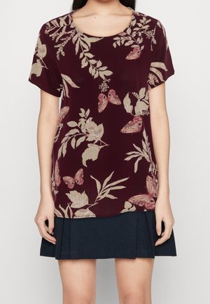Femme portant un haut à manches courtes bordeaux avec des motifs floraux et papillons beiges, associé à une jupe plissée foncée.