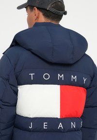 Person wearing a navy blue puffer jacket with a large white and red rectangular logo and "TOMMY JEANS" text on the back.