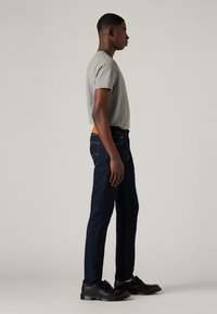 Joven de pie de lado, vistiendo una camiseta gris, jeans azul oscuro, calcetines blancos y zapatos negros con cordones, contra un fondo simple.