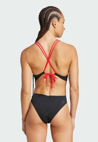 Černé bikini s červenými křížovými ramínky na zádech, s uzavíratelným zavazováním. Vysoké kalhotky s hladkým povrchem a minimálním logem.