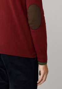Maglione rosso con orlo e polsini a coste, con toppe marroni sui gomiti. Indossato sopra una manica interna verde scuro. Materiale a maglia testurizzato.