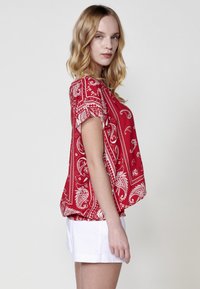 Top corto rosso a maniche corte con motivo paisley, vestibilità rilassata, scollo rotondo e tessuto leggero. Abbinato a pantaloni corti bianchi.