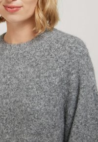 Pull en tricot gris fabriqué à partir d'un matériau doux et texturé. Il présente un col côtelé et une coupe décontractée, avec un motif chiné subtil sur l'ensemble.